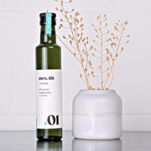 Kaltgepresstes Leindotteröl von mrs. tüt Ölmanufaktur in 250-ml-Flasche, dekoriert mit Leindotterpflanze auf einem Edelstahltisch vor weißem Hintergrund