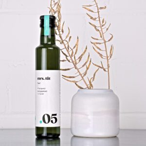 Kaltgepresstes Senföl von mrs. tüt Ölmanufaktur in 250-ml-Flasche, dekoriert mit Senfpflanze auf einem Edelstahltisch vor weißem Hintergrund