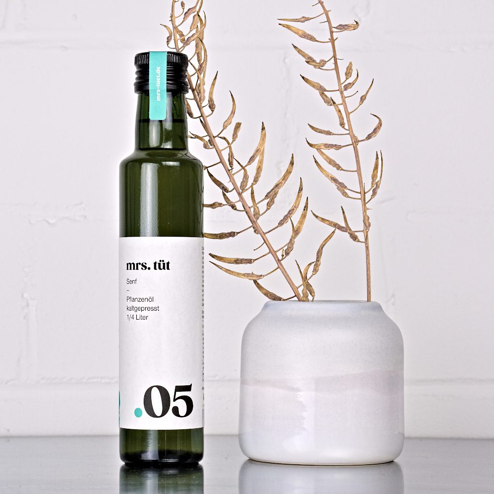Kaltgepresstes Senföl von mrs. tüt Ölmanufaktur in 250-ml-Flasche, dekoriert mit Senfpflanze auf einem Edelstahltisch vor weißem Hintergrund