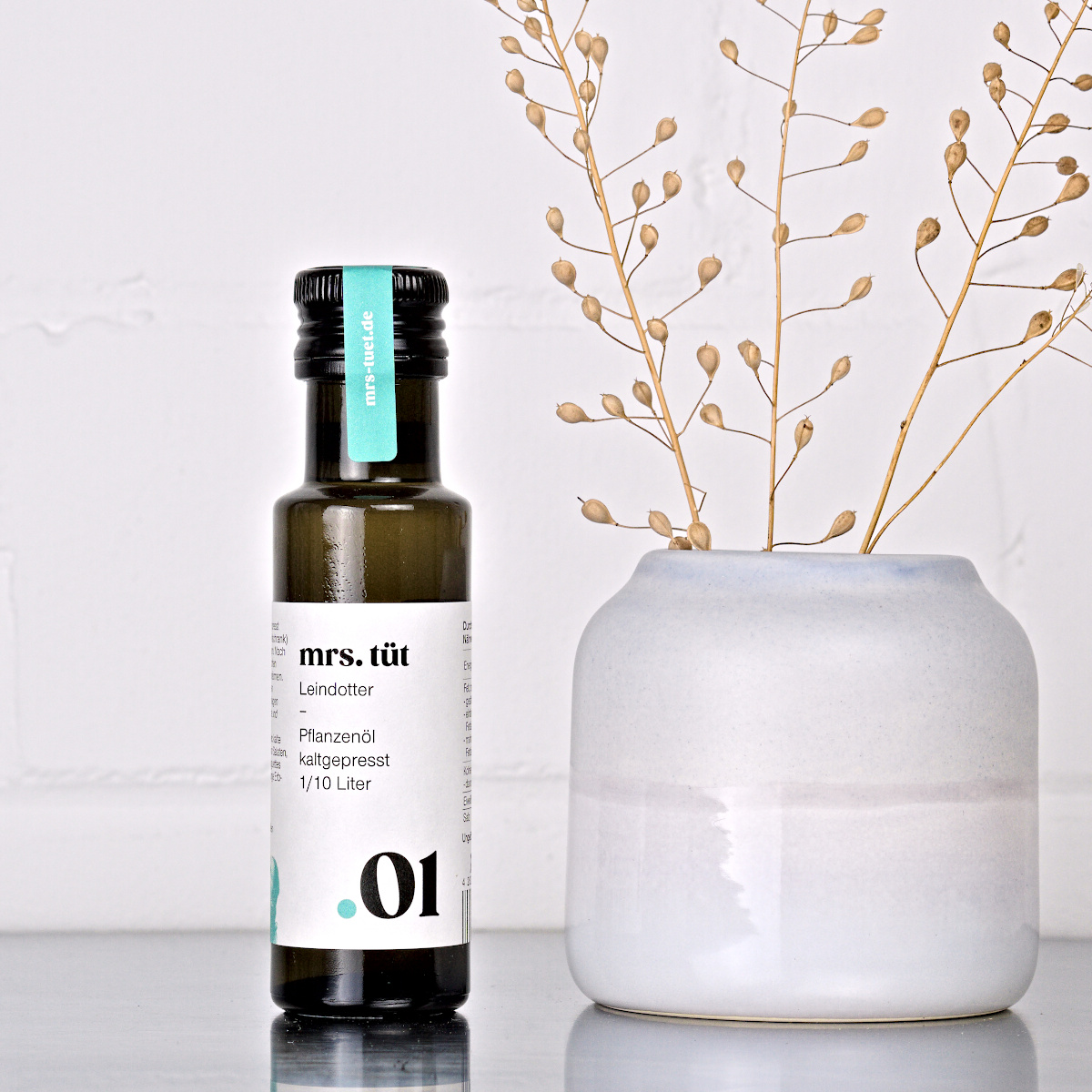 Kaltgepresstes Leindotteröl von mrs. tüt Ölmanufaktur in 100-ml-Flasche, dekoriert mit Leindotterpflanze auf einem Edelstahltisch vor weißem Hintergrund
