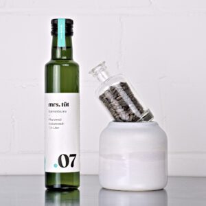 Kaltgepresstes Sonnenblumenöl von mrs. tüt Ölmanufaktur in 250-ml-Flasche, dekoriert mit Sonnenblumensaat auf einem Edelstahltisch vor weißem Hintergrund
