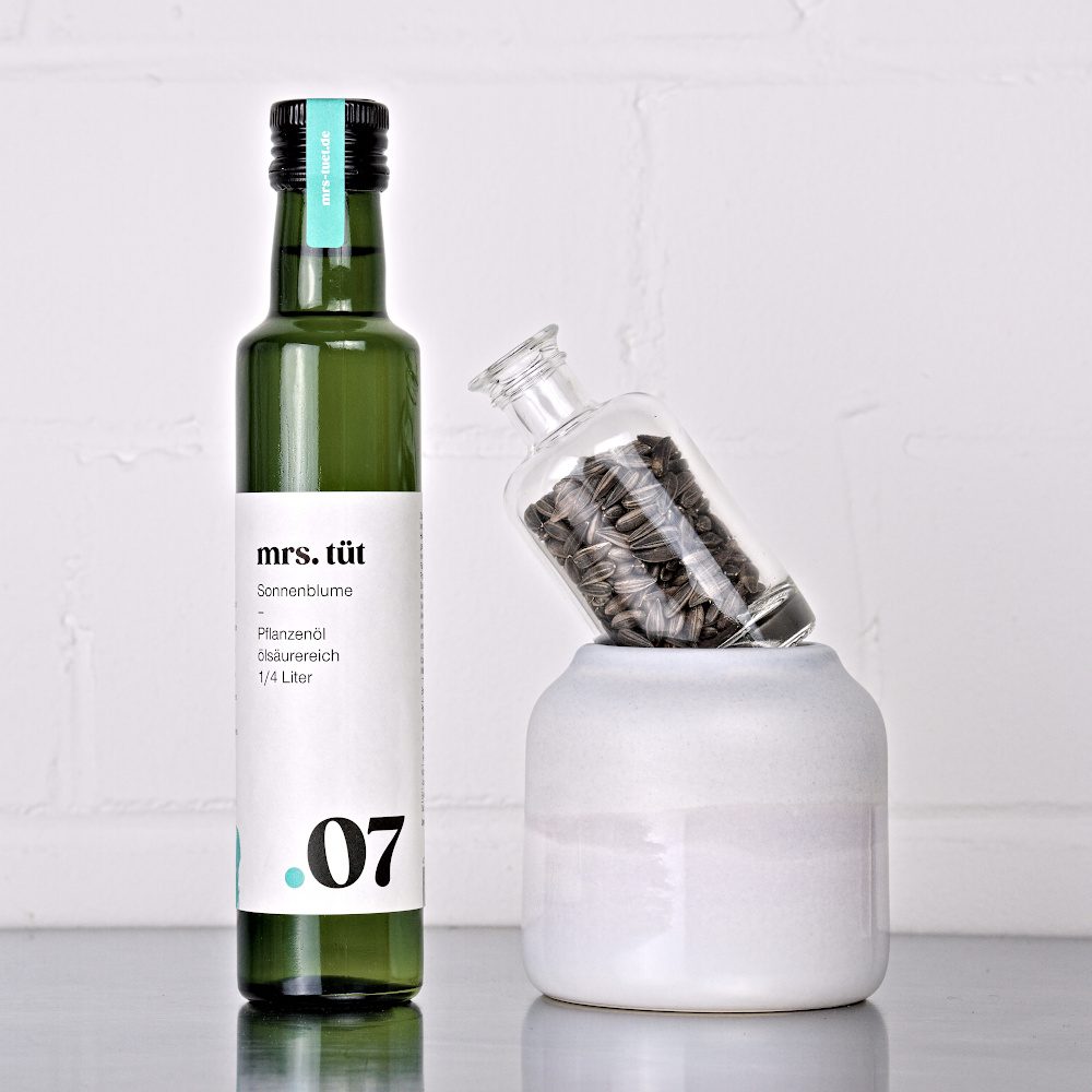 Kaltgepresstes Sonnenblumenöl von mrs. tüt Ölmanufaktur in 250-ml-Flasche, dekoriert mit Sonnenblumensaat auf einem Edelstahltisch vor weißem Hintergrund