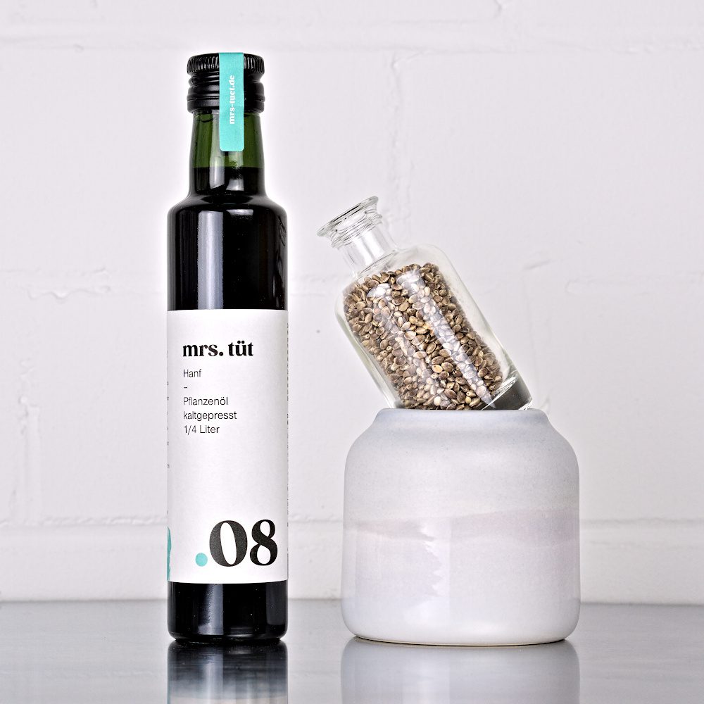 Kaltgepresstes Hanföl von mrs. tüt Ölmanufaktur in 250-ml-Flasche, dekoriert mit Hanfsaat auf einem Edelstahltisch vor weißem Hintergrund