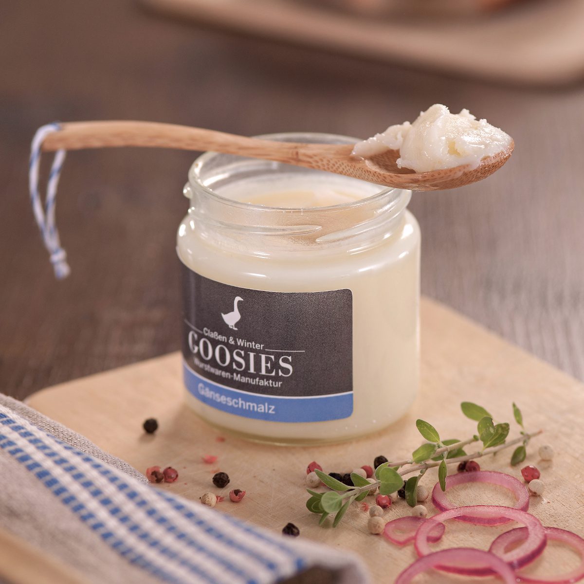 Reines Gänseschmalz der Goosies Wurstwarenmanufaktur im 175-g-Glas, dekoriert auf einem Holztisch mit Pfeffer und Zwiebeln