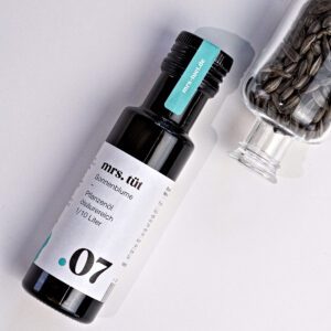 Kaltgepresstes Sonnenblumenöl von mrs. tüt Ölmanufaktur in 100-ml-Flasche, dekoriert mit Sonnenblumensamen im Glas vor einem weißem Hintergrund