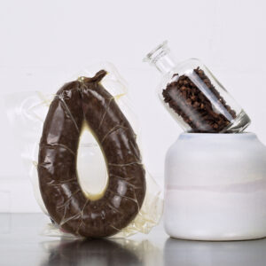 Rotwurst im Ring von der Gans der Goosies Wurstwarenmanufaktur in einer 225-g-Packung, dekoriert mit Nelken auf einem Edelstahltisch vor wei&szlig;em Hintergrund