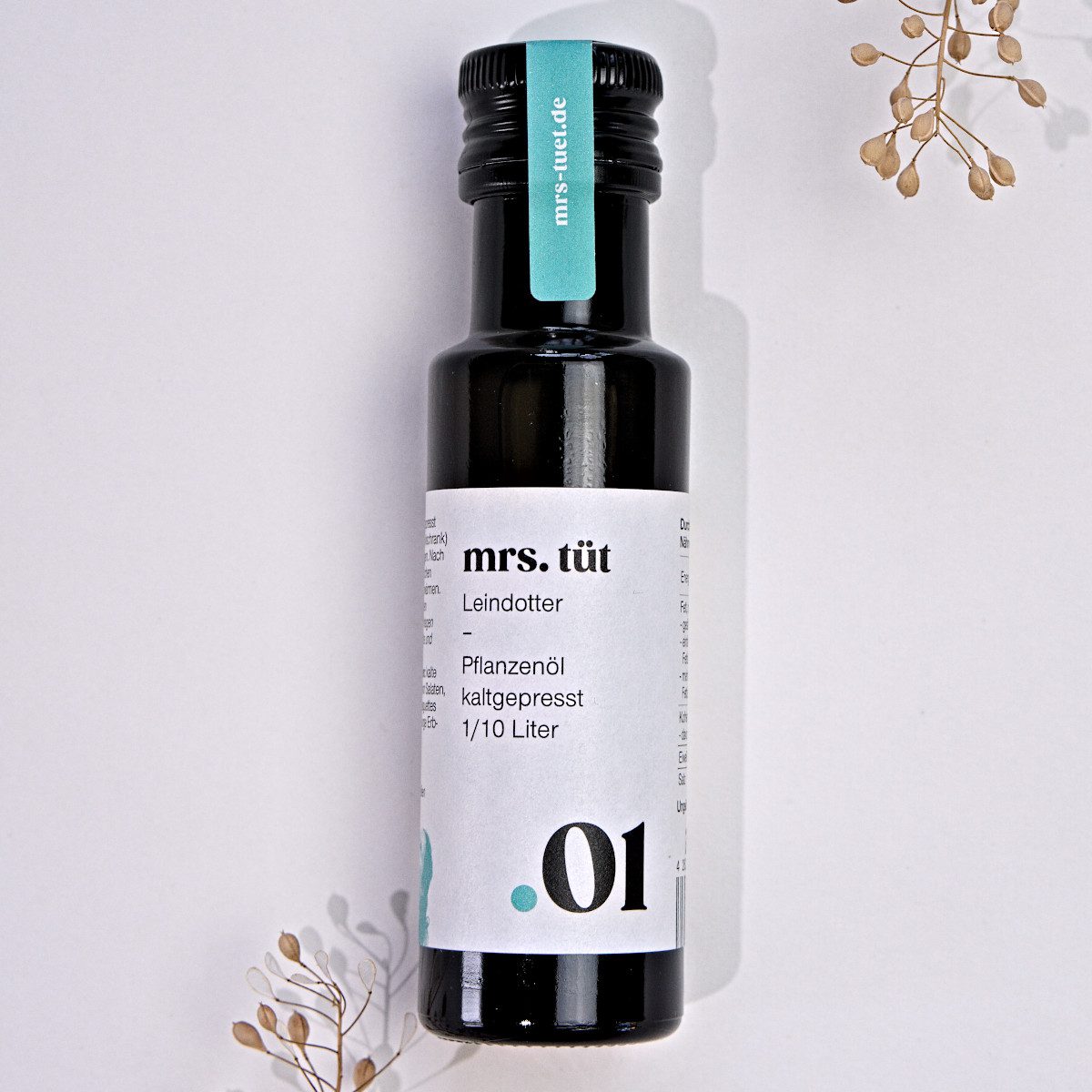 Kaltgepresstes Leindotteröl von mrs. tüt Ölmanufaktur in 100-ml-Flasche, dekoriert mit Leindotterpflanze vor einem weißem Hintergrund